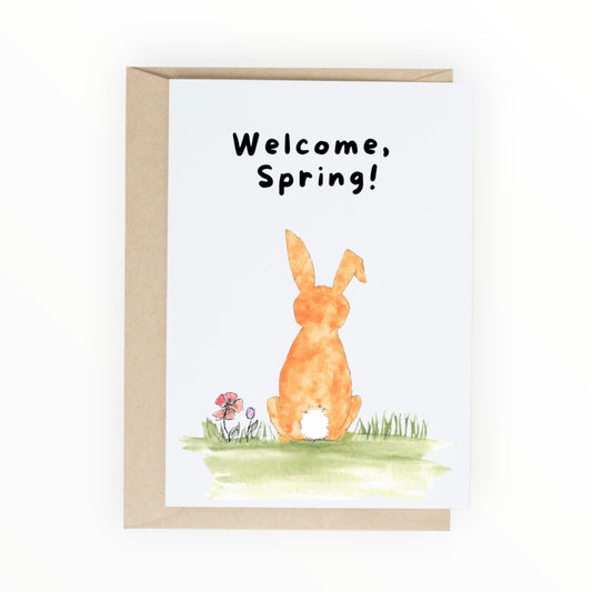 Orange watercolor bunny sitting in grass and flowers with text ‘Welcome, Spring.